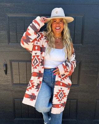 A woman stands in front of a black garage door wearing a cream and rust Southwestern patterned cardigan, a white tank top, blue jeans, a beige wide-brim hat, and gray boots.