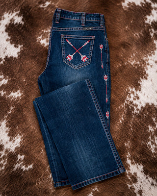 A pair of dark blue denim jeans laid flat on a brown and white cowhide rug. The back pocket features embroidered red and white floral and arrow details, and matching embroidery runs down the side seam of the leg.