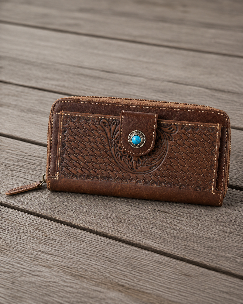 A brown leather zip-around wallet with embossed western-style floral and basketweave patterns, featuring a snap-button closure with a turquoise accent, resting on a weathered wooden plank surface.