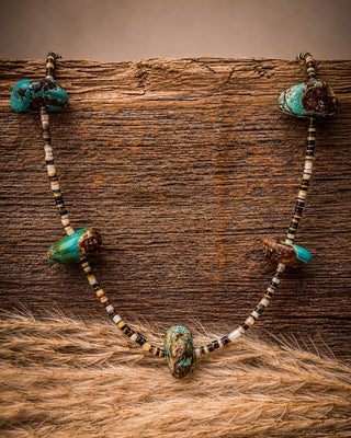 A beaded necklace with turquoise-colored stone beads and small black-and-white spacer beads, displayed draped over a rustic wooden surface with tan fur in the foreground.