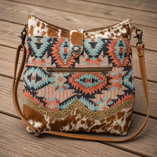A western-style bucket shoulder bag with a brown and white cowhide pattern, a turquoise and coral geometric woven front panel, and tan leather straps, shown on a wooden deck.