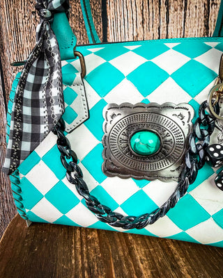A turquoise and white checkered handbag with a western-style silver concho featuring a turquoise stone, a black and white checkered scarf, and a braided black and turquoise shoulder strap, set against a rustic wooden background.