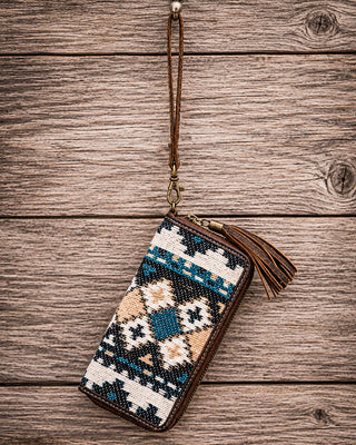 A zip-around wallet with a Southwestern geometric pattern in blue, beige, and white, featuring dark brown leather trim, a wrist strap, and a tassel, displayed on a rustic wooden background.