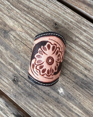 A handmade leather scarf slide with a floral tooling design, resting on a rustic wooden surface.