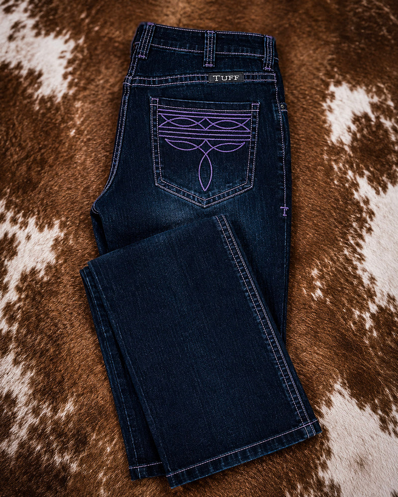 A pair of dark blue denim jeans laid flat on a brown and white cowhide rug. The back pocket features purple embroidered geometric and leaf-like patterns, and a small 'Tuft' brand label is visible at the back waistband.