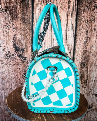 A turquoise and white checkered handbag with a pearl chain strap, displayed on a round wooden stool against a rustic wood plank background.