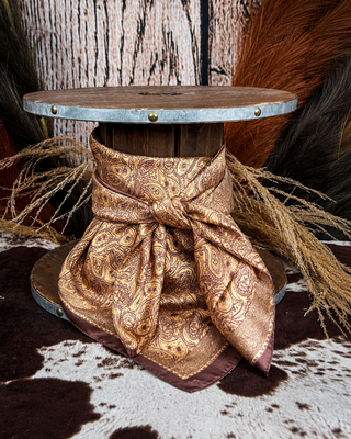 A square silk scarf with a brown paisley pattern and a dark brown border, tied in a large bow around the base of a rustic wooden spool. The spool is styled with dried grasses, a cowhide rug, and a wood-paneled background.