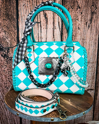A turquoise and white harlequin-patterned handbag with a matching detachable shoulder strap, displayed on a rustic wooden stool against a wood plank backdrop.