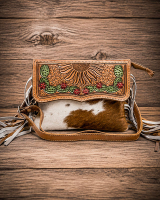 A hand-tooled leather clutch bag with a brown and white cowhide body, featuring a sunflower and cactus design on the flap, set against a rustic wooden background.