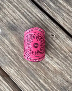 A bright pink leather scarf slide with an embossed floral pattern, resting on weathered gray wooden planks.