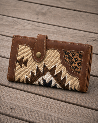A brown leather and woven textile wallet with a Southwestern-style geometric pattern, resting on a gray wooden plank surface.