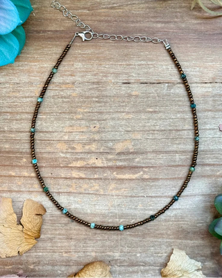 A beaded necklace with dark brown beads, small turquoise blue beads, and silver-toned chain extender, displayed on a rustic wooden surface with dried flowers and succulents.