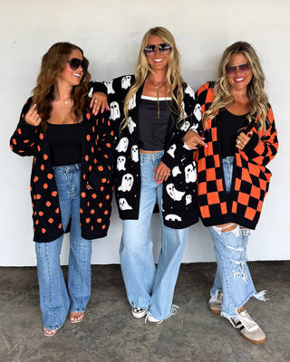 Three women wearing colorful cardigans and jeans standing against a white wall.