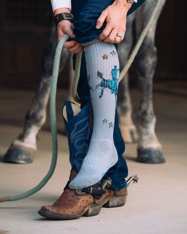 Model posing in the Long Live Grey Western Cowboy Performance Socks with a horse in the back ground.