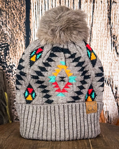 The Aztec Pom Beanie displayed on a wooden table with a wood background.