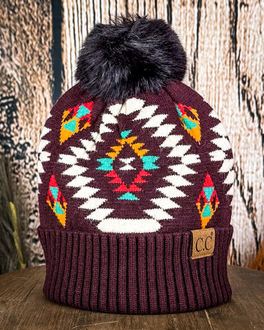 The Aztec Pom Beanie displayed on a wooden table with a wood background.