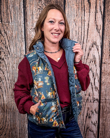 A girl modeling the Jolie Hooey Reversible Puffer Vest. The vest is open, she is wearing a red sweater underneath. She has one hand in her pocket and is holding the collar of the vest in her other hand. She is standing in front of a wood backdrop.