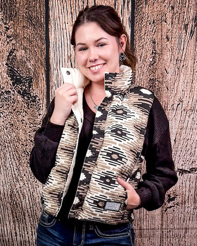 A girl modeling the Jane Hooey Reversible Puffer Vest with one hand in her pocket and the other is holding the collar of the vest. The vest is open, showing a sweater underneath. There is a wood backdrop behind her.