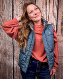 A girl modeling the Jolie Hooey Reversible Puffer Vest. The vest is open and inside out to show the reversable design. She has one hand by her leg and the other in her hair. She is standing in front of a wood backdrop.
