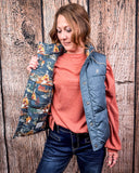 A girl wearing the Jolie Hooey Reversible Puffer Vest. The vest is inside out to show the reversable design. She is holding the vest open to show the print on the inside. She is standing in front of a wood backdrop.
