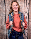 A girl modeling the Jolie Hooey Reversible Puffer Vest. The vest is inside out to show the reversable design. She is holding the sides of the vest. She is standing in front of a wood backdrop. 