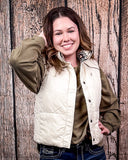 A girl modeling the Jane Hooey Reversible Puffer Vest. It is turned inside out to show the reversable design. She has one hand in her pocket and one hand in her hair. She is standing in front of a wood backdrop.