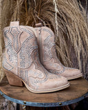Pair of beige cowboy boots with rhinestone details on a wooden surface with dried grass in the background.