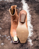 Brown woven ankle boot with gold tip on a textured brown surface