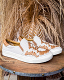 White slip-on sneakers with brown accents on a wooden surface with decorative background