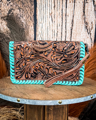 Handbag with turquoise edge and intricate brown leather design on a wooden surface.