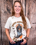 Woman wearing a white t-shirt with a graphic design of a boot, moon, and text against a wooden background.