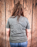 Person wearing a gray shirt and blue jeans standing against a wooden wall.