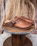 Brown faux suede shoes with buckles on a wooden spool against a rustic background