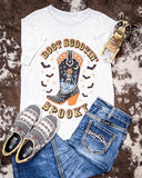 White t-shirt with 'Boot Scootin' Spooky' design, blue jeans, and patterned shoes on a textured surface.