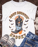 White t-shirt with Halloween-themed design featuring a boot, skeleton, and pumpkin, paired with jeans and slippers on a textured surface.