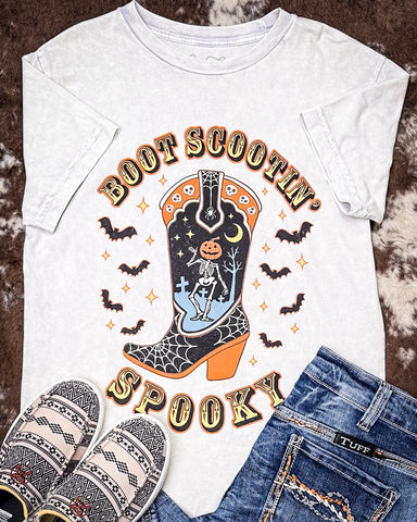 White t-shirt with Halloween-themed design featuring a boot, skeleton, and pumpkin, paired with jeans and slippers on a textured surface.