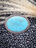 Western-style turquoise belt buckle with silver floral trim displayed on black beans and dried grass