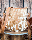 A brown and white hair-on-hide crossbody bag with a leather strap and fringe, displayed on a wooden surface.