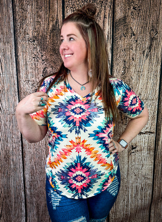 A woman wearing a colorful short-sleeve V-neck top with a vibrant geometric and tribal-inspired pattern in pink, orange, blue, and yellow on a cream background, paired with distressed blue jeans, posing against a rustic wooden plank wall.
