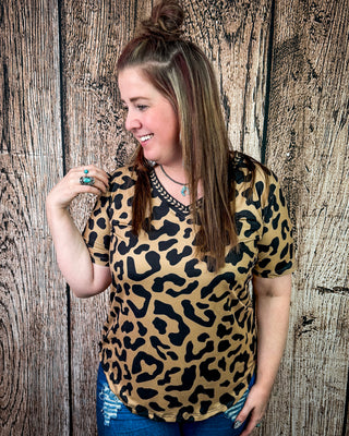 A woman wearing a leopard print short-sleeve top with a v-neck and black lace trim, standing against a rustic wood plank wall.