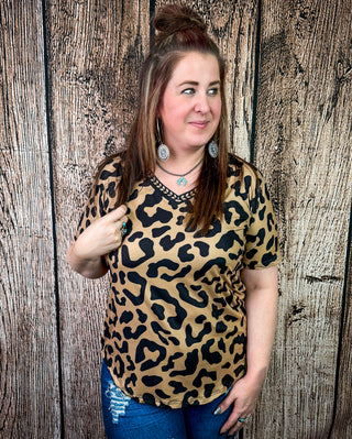 A woman wearing a tan and black leopard print short-sleeve top with a black embroidered V-neck trim, posing in front of a rustic wooden plank wall.