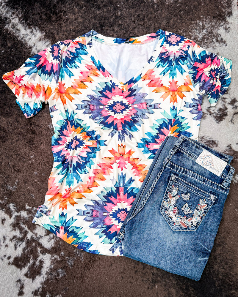 A short-sleeve V-neck top with a colorful geometric starburst pattern in pink, blue, orange, and purple on a white base, styled flat-lay with folded blue jeans on a brown and white faux fur surface.