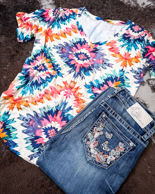 A short-sleeve V-neck top with a colorful geometric starburst pattern in blue, pink, orange, and purple on a white base, styled flat-lay next to a pair of blue jeans with embroidered back pocket details on a dark brown faux fur surface.