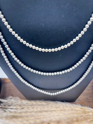 Three strands of silver beaded necklaces draped on a black jewelry display bust, with a soft beige textured fabric in the foreground.