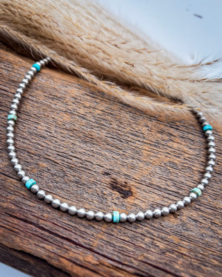 A beaded necklace with small silver-toned round beads and turquoise-colored spacer accents, displayed on a rustic wooden surface with dried grass in the background.