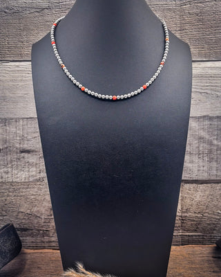 A beaded necklace displayed on a black mannequin bust against a rustic wood plank background. The necklace features small silver-gray beads with alternating coral-red and dark gray bead accents.