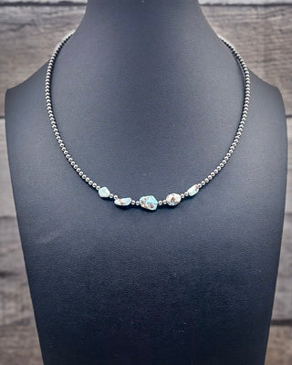 A silver beaded necklace with five iridescent, irregularly shaped blue-green stones, displayed on a black jewelry bust against a rustic gray wood background.