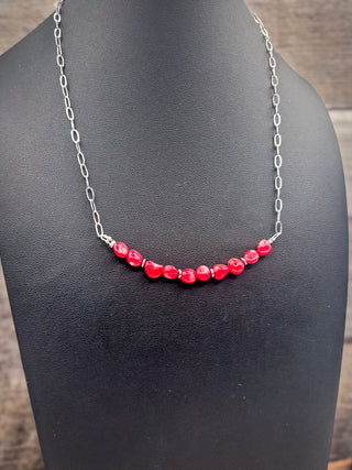 A silver chain necklace featuring a curved row of faceted red beads, displayed on a black jewelry bust against a dark wood background.
