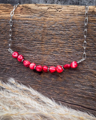 A silver chain necklace featuring a curved row of glossy, irregular red beads, displayed on a rustic dark wood background with dried pampas grass in the foreground.