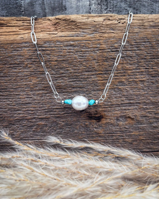 A silver paperclip chain necklace featuring a central white pearl flanked by two small turquoise beads, displayed on a rustic wooden surface with dried grass in the foreground.